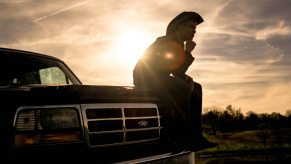 Man in a cowboy hat sitting on the hood of a black Ford F-250 truck at sunset.