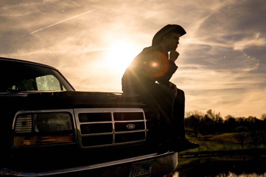 Man in a cowboy hat sitting on the hood of a black Ford F-250 truck at sunset.