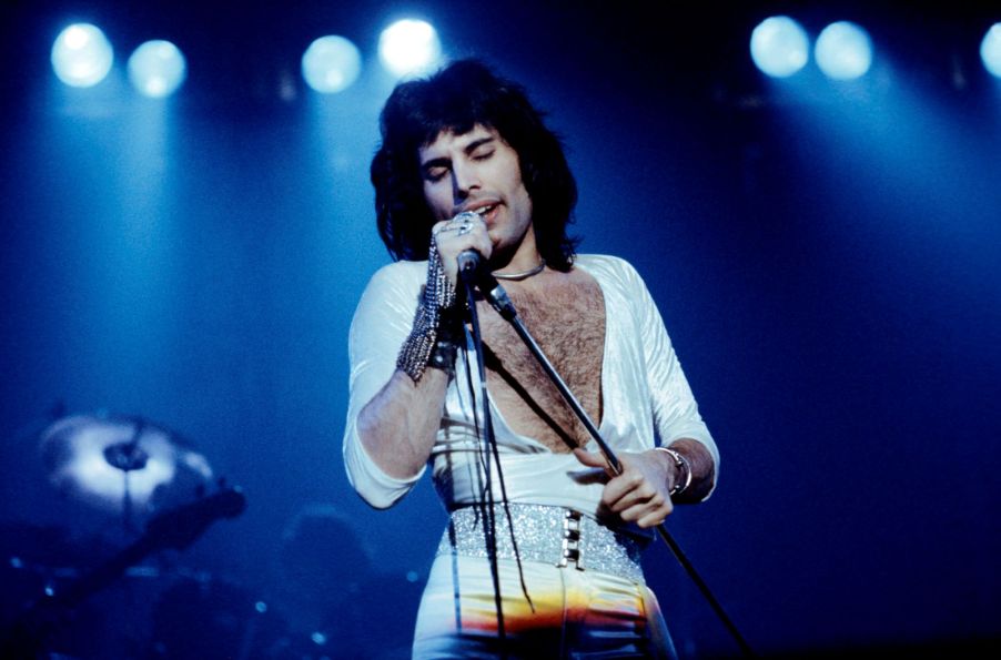 Freddie Mercury from Queen singing on stage in a white open-chest shirt and white pants with a silver glittery belt with black buttons with lights in the background.