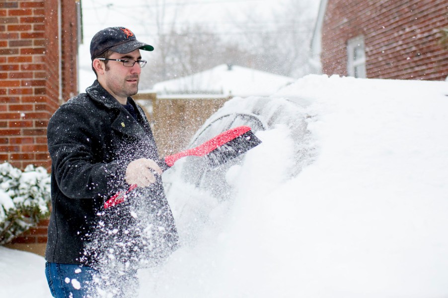 A man in a baseball cap stands in front of a brick house and uses a tool to brush the snow off a car.