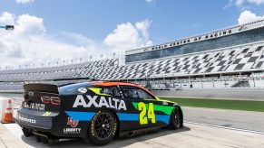 A NASCAR race car parked on the edge of the Daytona Speedway, the stands visible in the distance.