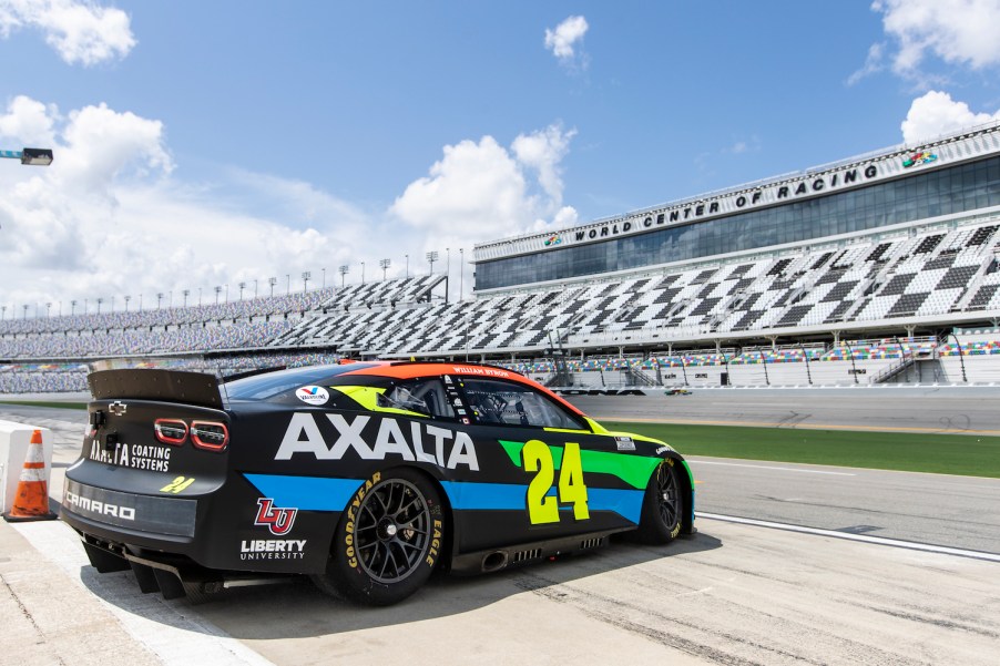 A NASCAR race car parked on the edge of the Daytona Speedway, the stands visible in the distance.