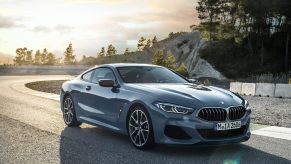 A 3/4 front view of a blue/grey 2022 BMW 8 Series Coupe parked at a corner of a race track. The background shows trees and rock cliffs.