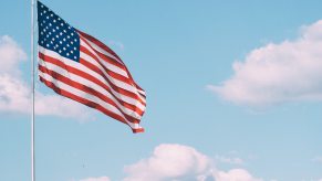 A large American flag blowing in the breeze in front of a light blue sky and fluffy white clouds.