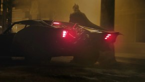 Robert Pattinson looks into the distance while standing next to his muscle car batmobile.