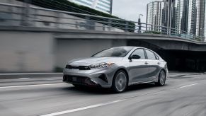 The 2022 Kia Forte compact sedan in gray driving past a concrete bridge in an urban city