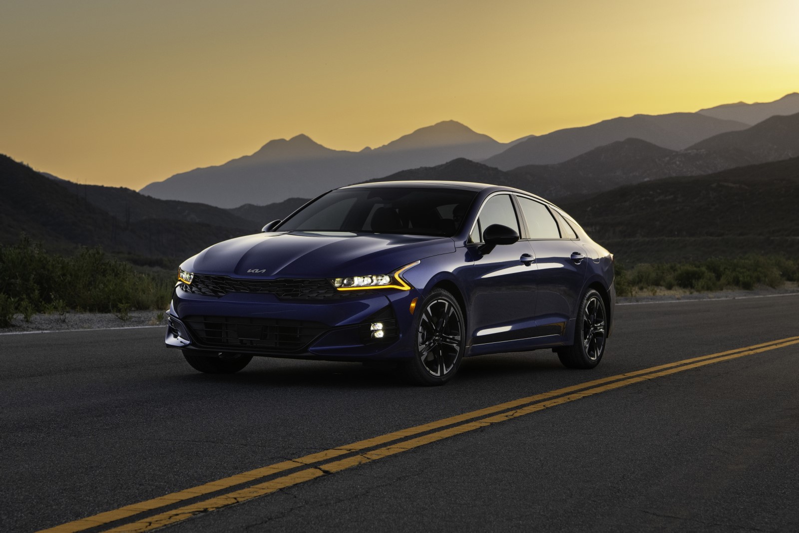 Angled front view of the 2022 Kia K5 in dark blue, driving on a road at dusk with mountains in the background