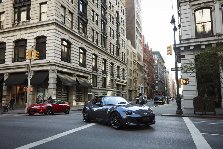 A front view of a grey 2022 Mazda Miata RF and a red Miata convertible on a city street with buildings in the background.