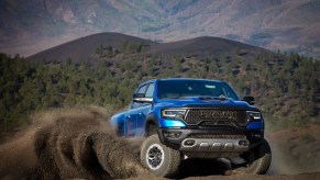 A blue Ram 1500 pickup truck showing off its TRX Hellcat supertruck engine by skidding across the dirt, with wooded hills in the background.