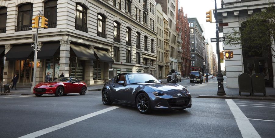 A front view of a grey 2022 Mazda Miata RF and a red Miata convertible on a city street with buildings in the background.