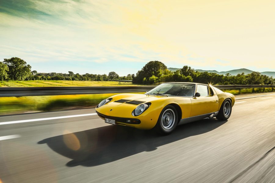 A 3/4 front view of a yellow Lamborghini Miura driving on a road with trees and hills in the background.