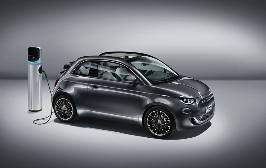 A profile view of a grey 2022 Fiat 500e plugged into an EV charging station. Car is shown with a grey studio background.