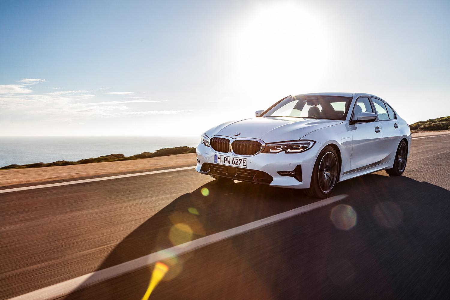 White 2022 BMW 330e plug-in hybrid sedan driving down coastal highway at sunset