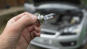 A hand holding a headlight replacement bulb in front of a car with its hood up.