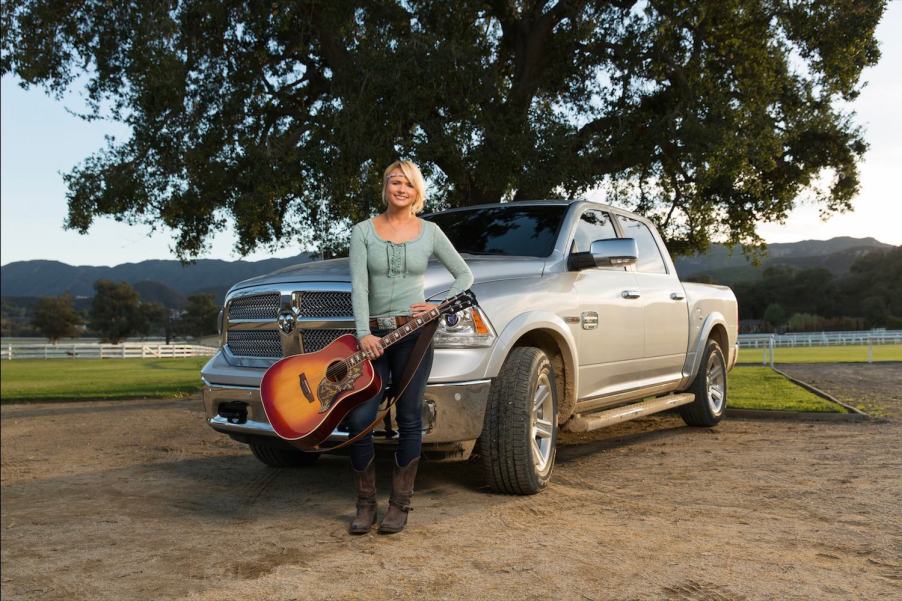 Woman with a guitar leans against a Ram 1500 parked on a farm, in front of a tree.