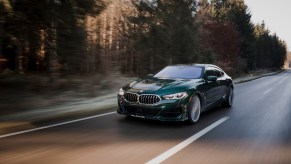 A 3/4 front view of a green Alpina B8 sedan driving on a tree-lined road.