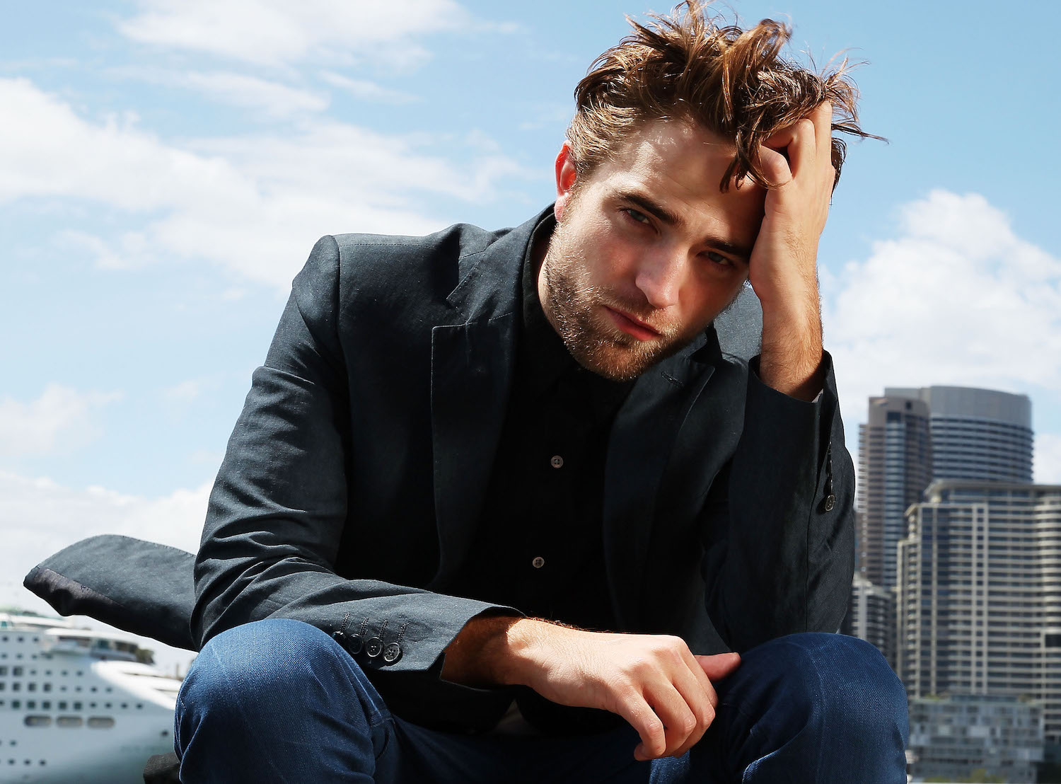 Publicity shot of actor Robert Pattinson dressed in jeans and a suit jacket, the daytime skyline of a city visible behind him.