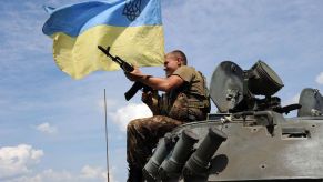 Ukrainian military soldier holds a machine gune and smiles while sitting atop an armored vehicle flying a Ukraine flag.