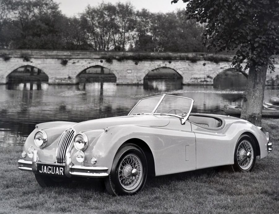 A white 1954 Jaguar XK140 Roadster parked next to a river bridge