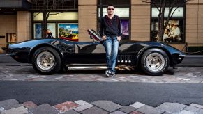 A Japanese business owner, wearing a black sweatshirt, jeans, and black sneakers, leans against his black 1969 Chevrolet Corvette