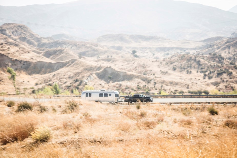 A pickup truck in the distance pulls an airstream trailer through the mountains, sandy ridgelines visible behind it.