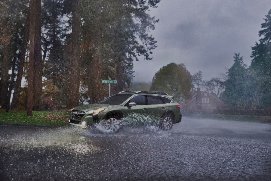 A green updated 2023 Subaru Outback splashing through a puddle as it makes a sharp turn in the rain.