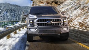 A gray 2022 Ford F-150 Platinum full-size pickup truck is driving on the road with snowy mountains in the background.