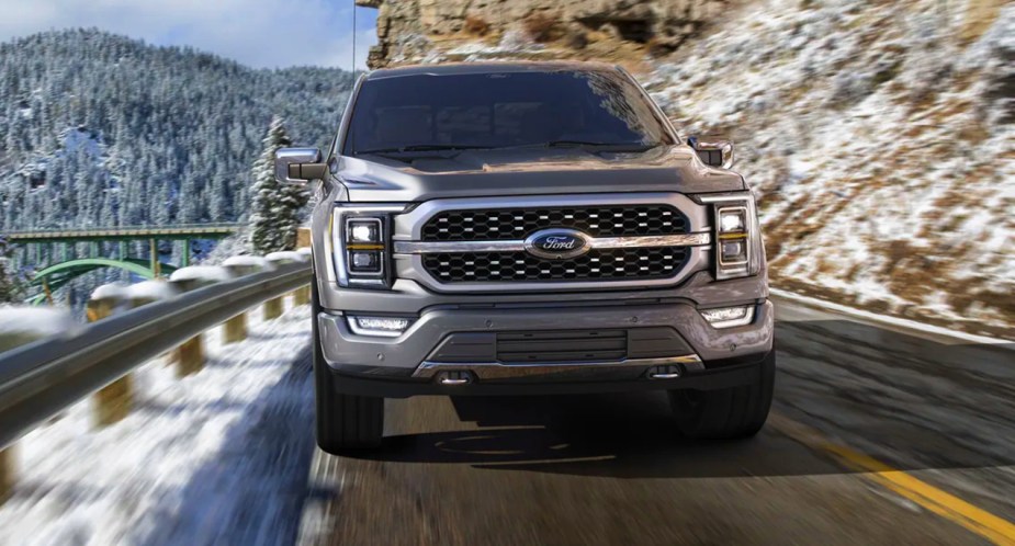 A gray 2022 Ford F-150 Platinum full-size pickup truck is driving on the road with snowy mountains in the background.