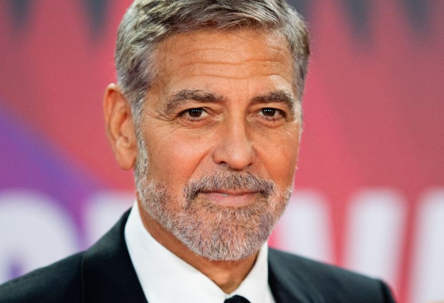 George Clooney standing in front of a pink and white background that is blurred.