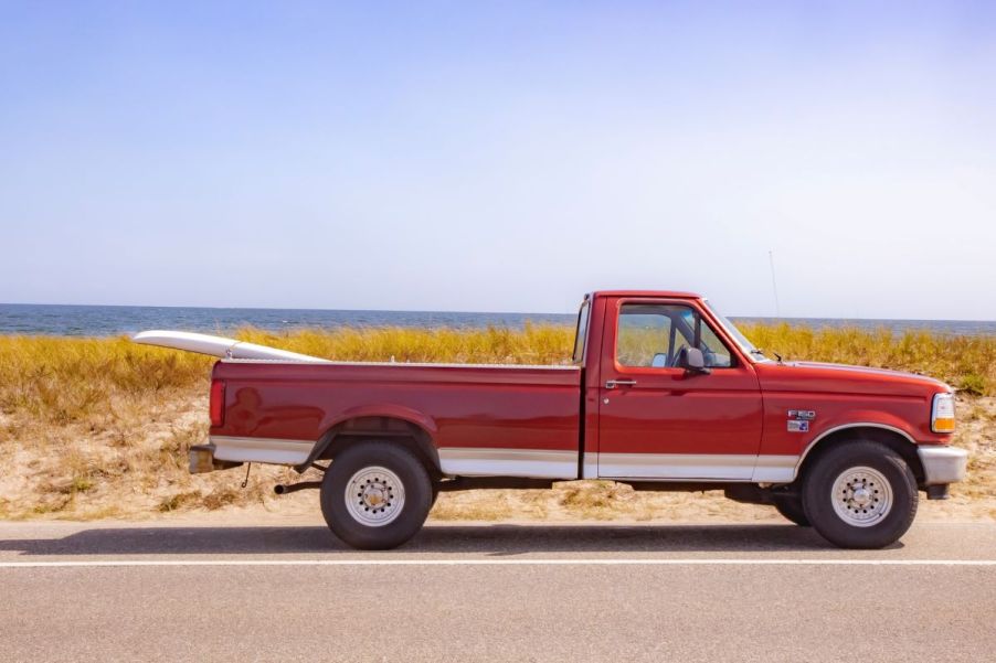 Well-maintained, classic Ford F-150 small pickup truck in red, parked in front of a beach with a surfboard strapped into the bed