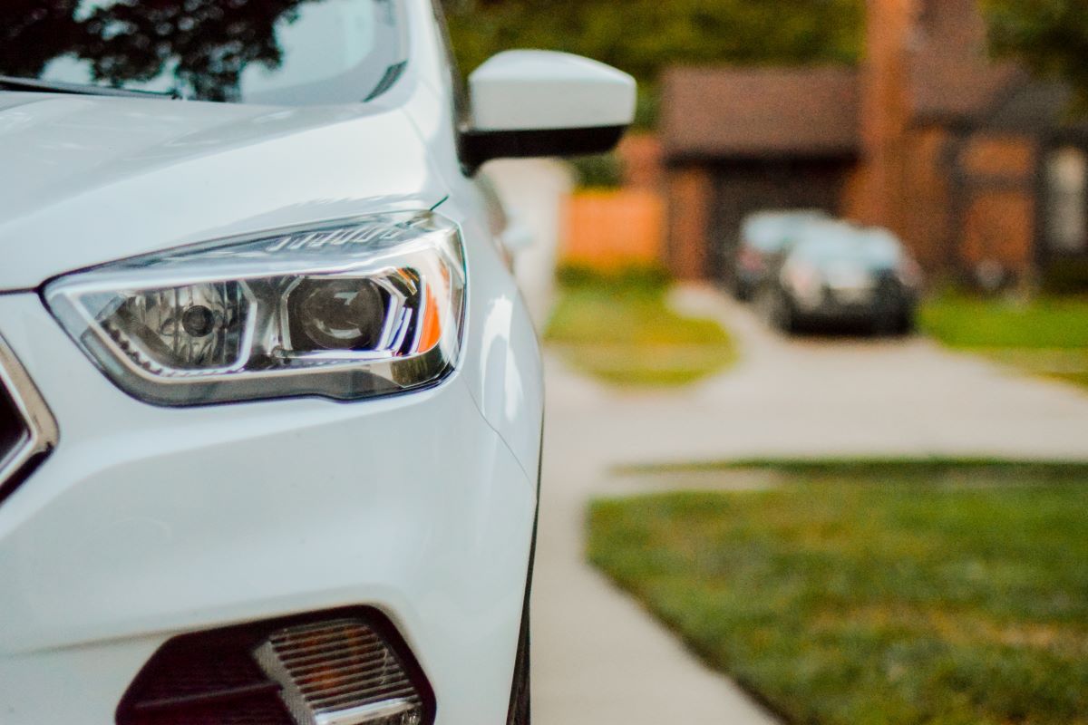 Close shot of a vehicle's front left headlight. Blurred background showing other cars parked in a driveway across the street