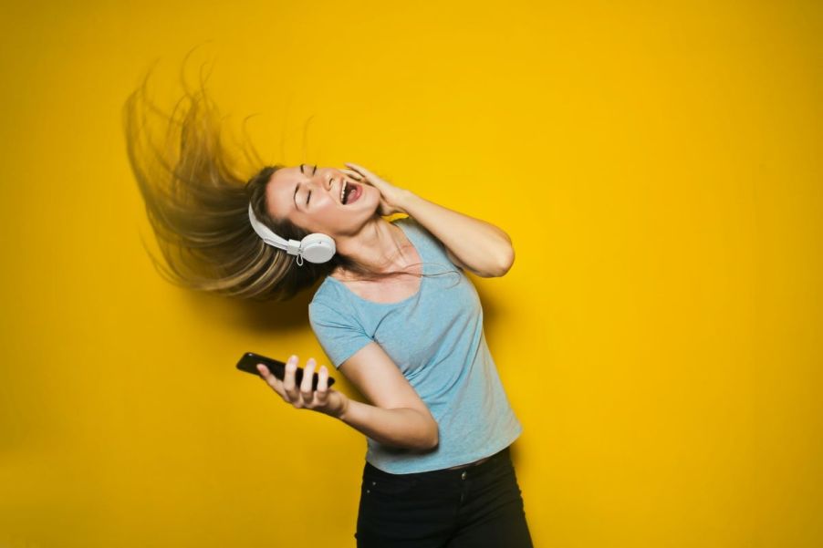 Blonde woman listening to upbeat music, dancing and whipping her hair. She's wearing white headphones, a green shirt, and black pants. She holds a smartphone in her right hand.