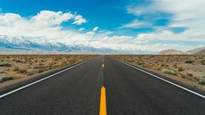 A beautiful long, open stretch of well-paved highway cutting through a plain. There are tall snow-capped mountains to the left and a blue sky with soft white clouds