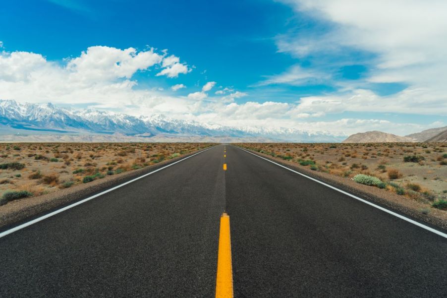 A beautiful long, open stretch of well-paved highway cutting through a plain. There are tall snow-capped mountains to the left and a blue sky with soft white clouds