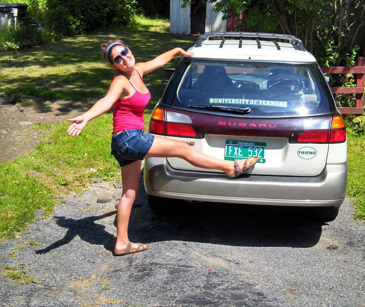 A young woman poses next to an old Subaru Outback, her first car; she would later sell it to a junkyard