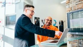 Two men stand together looking at a laptop inside of a business; 0% APR car financing is an attractive option for shoppers