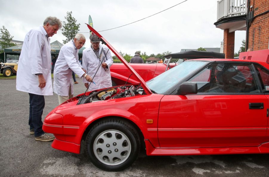 A group of Vintage Sports Car Club (VSSC) judges inspecting under the hood of a red 1998 Toyota MR2 Mk1