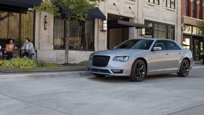 A 2022 Chrysler 300 with a light gray paint color parked on a concrete road near a shopping street and cafe