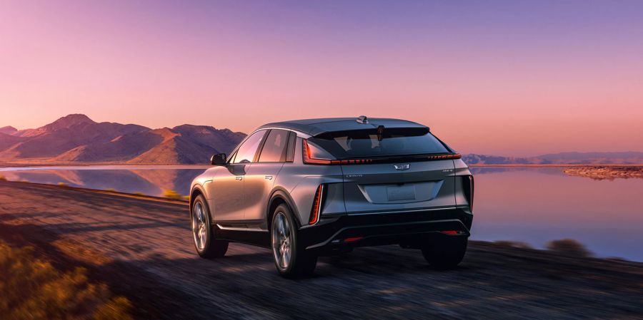 A rearview shot of a gray 2023 Cadillac Lyriq luxury electric SUV model driving near a lake at sunset