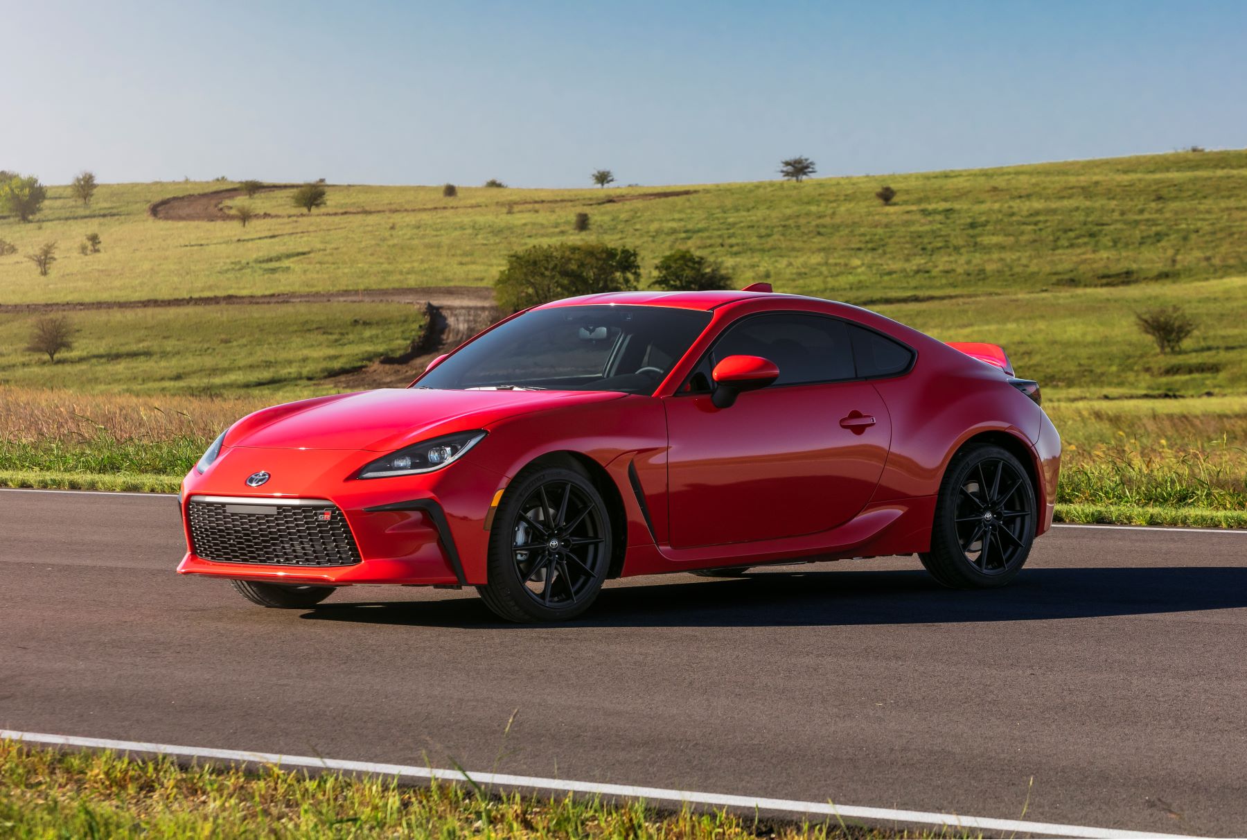 A red 2022 Toyota GR86 sports car parked on a country highway road near dirt paths dug into grass hills