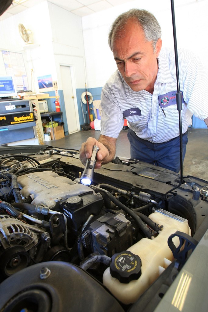 A technician visually inspects a car engine bay with a flashlight as part of an emissions test in California