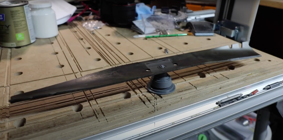 A sharpened lawn mower blade balances on a work bench, extra tools visible behind it.