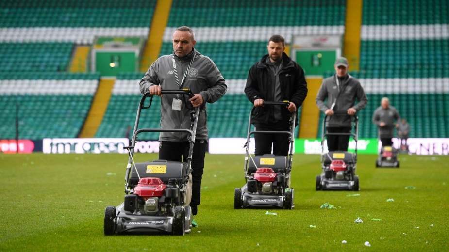 Individuals pushing lawn mowers across a sports field.