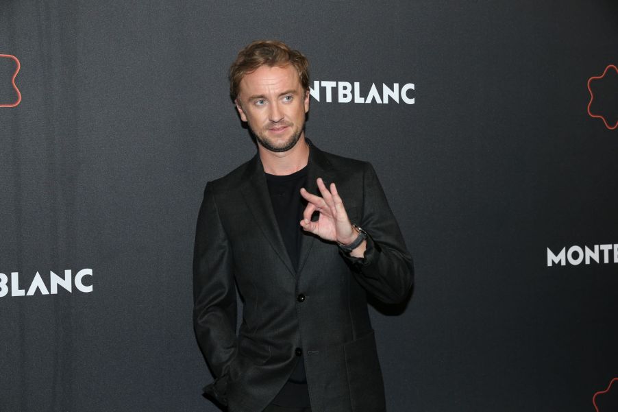 Tom Felton wearing a black suit with a black background.