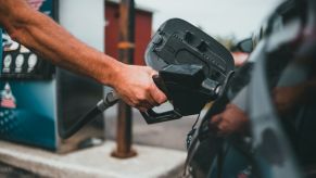 Muscular arm holding a gas pump nozzle up to the fuel cap of a car at a gas station. The best fuel rewards programs of 2022 can help you save money.