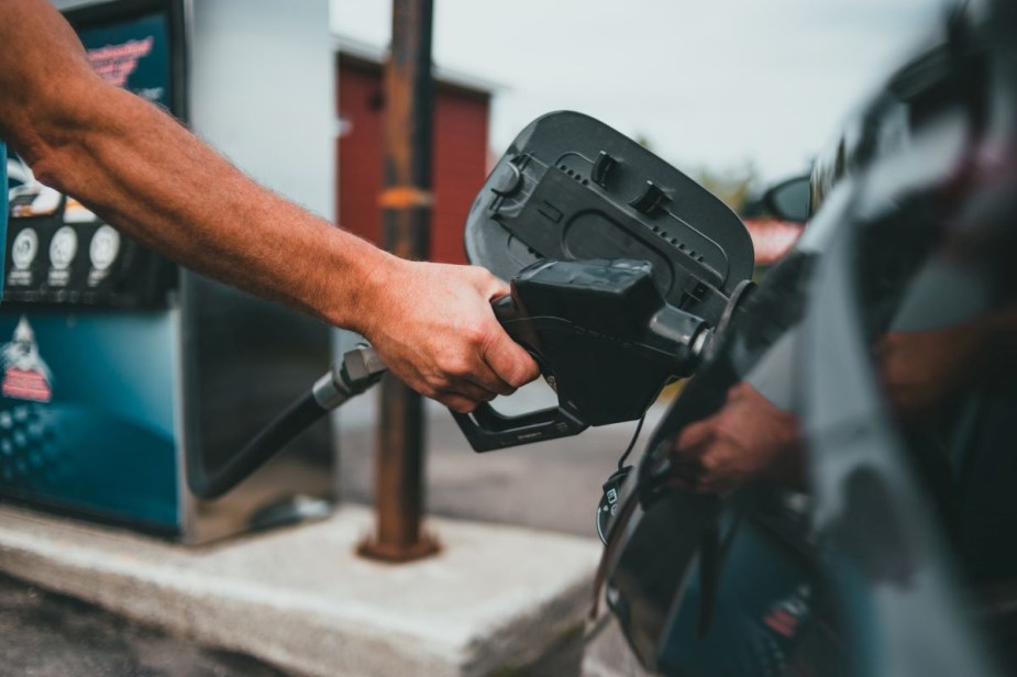 Muscular arm holding a gas pump nozzle up to the fuel cap of a car at a gas station. The best fuel rewards programs of 2022 can help you save money.