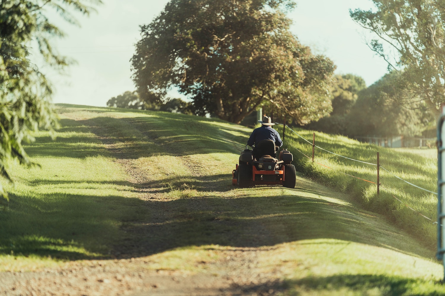 Man mowing grass along a fenceline with trees.