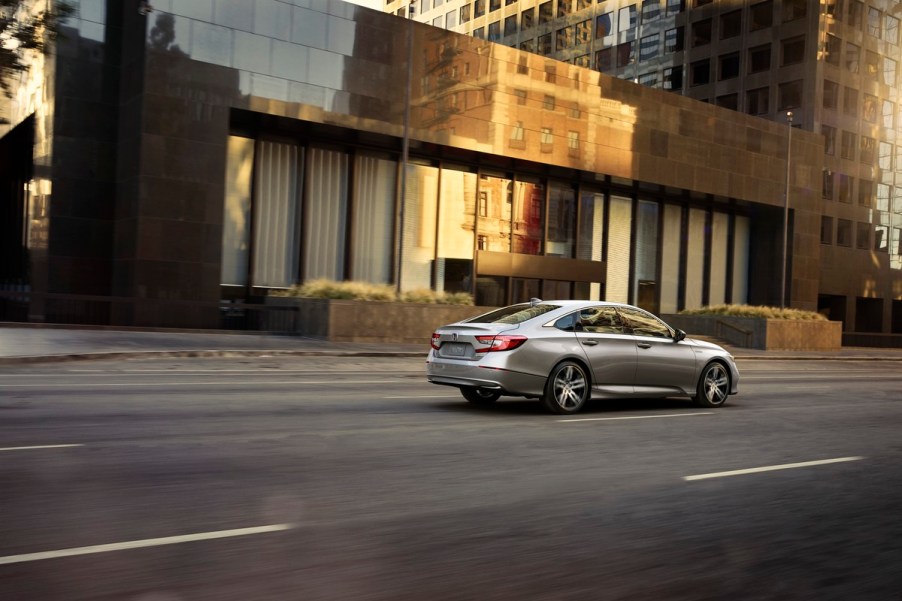 A silver 2021 Honda Accord Hybrid drives down a multi-lane road with a short commercial building in the background