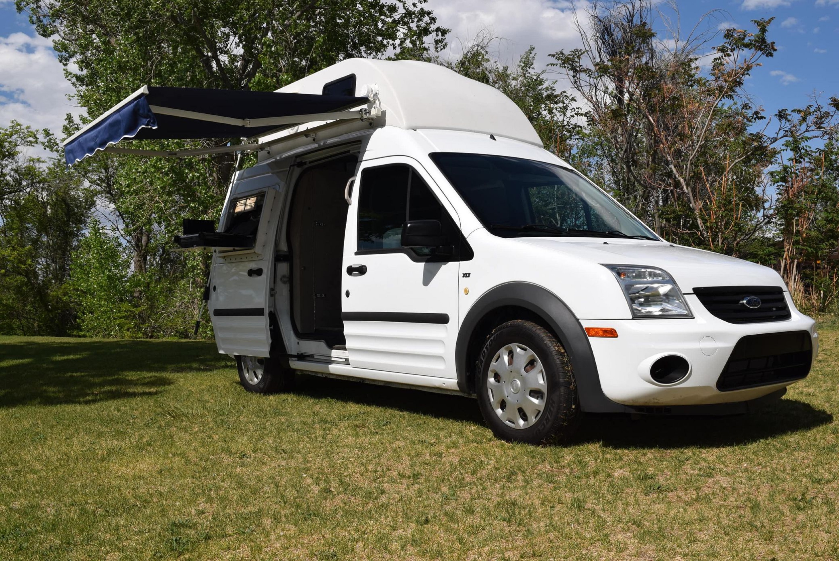 A white 2011 Ford Transit Connect XLT camper van in a field with its awning extended