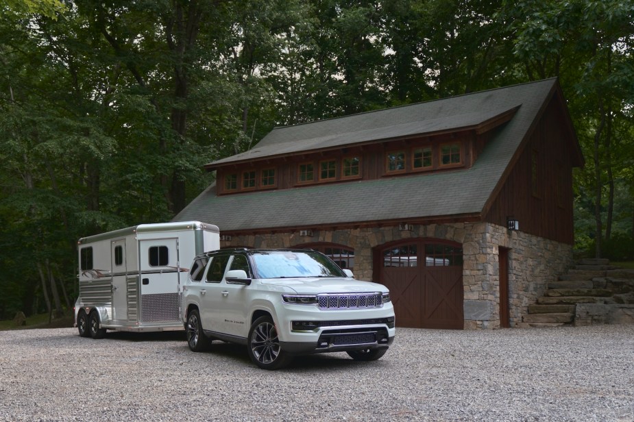 White Jeep Grand Wagoneer hooked up to a horse trailer, parked in front of a stable in the woods.